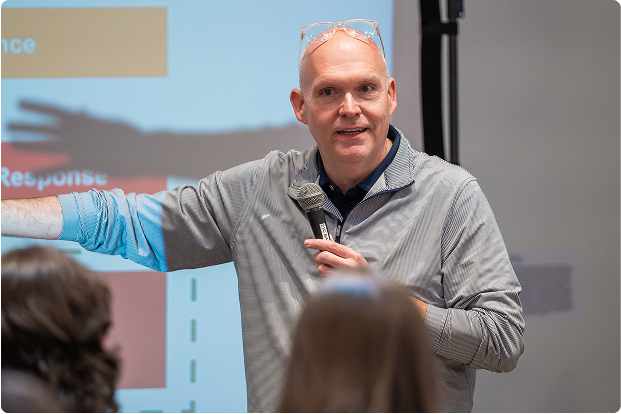 A man with glasses and a microphone presenting in front of a projector