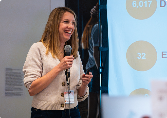 A smiling woman with a microphone presenting in front of a projector